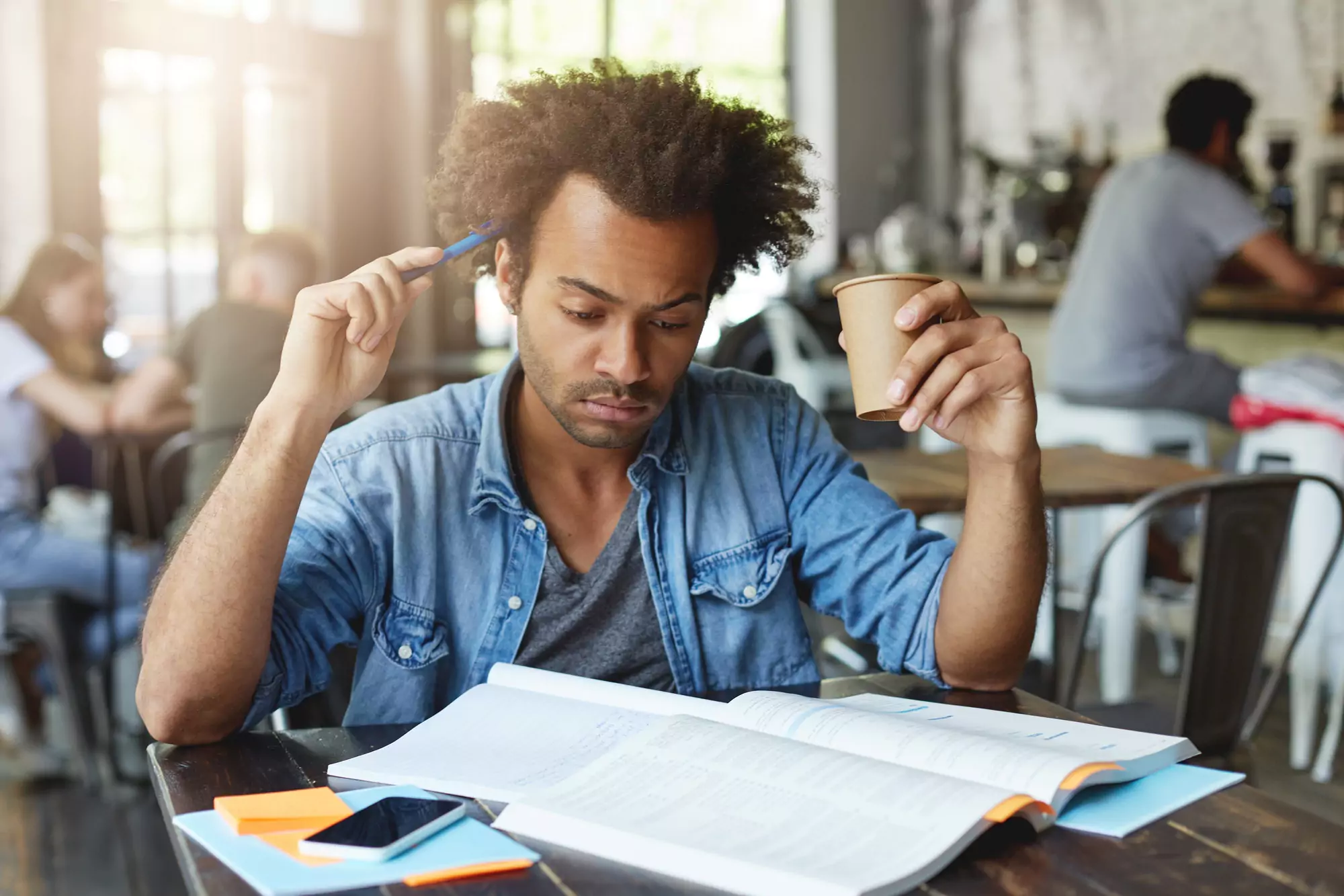 Concentrated thoughtful black European student with Afro hairstyle scratching head with pen, drinkin