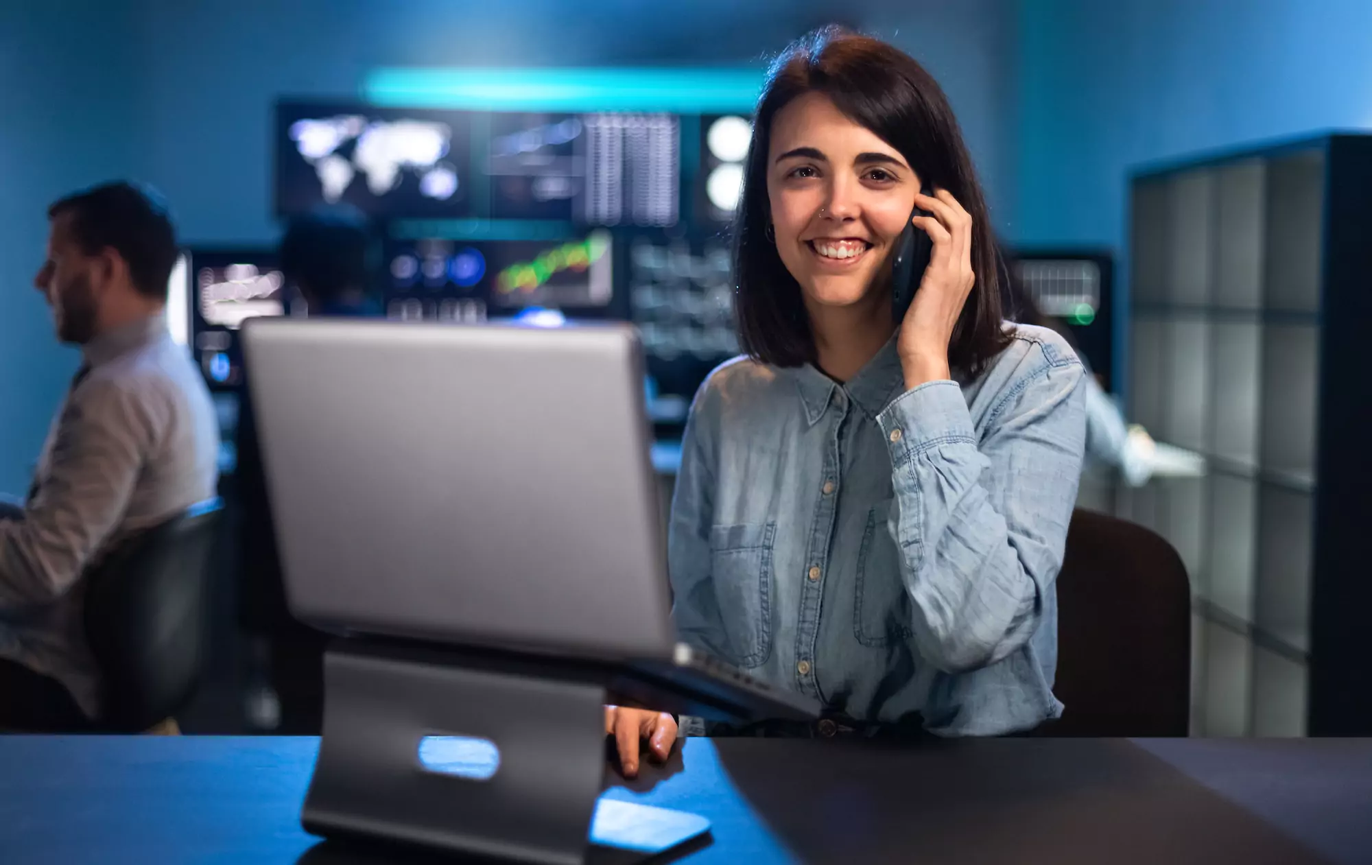 Female stock trader working in office using laptop and talking on the phone looking at camera.