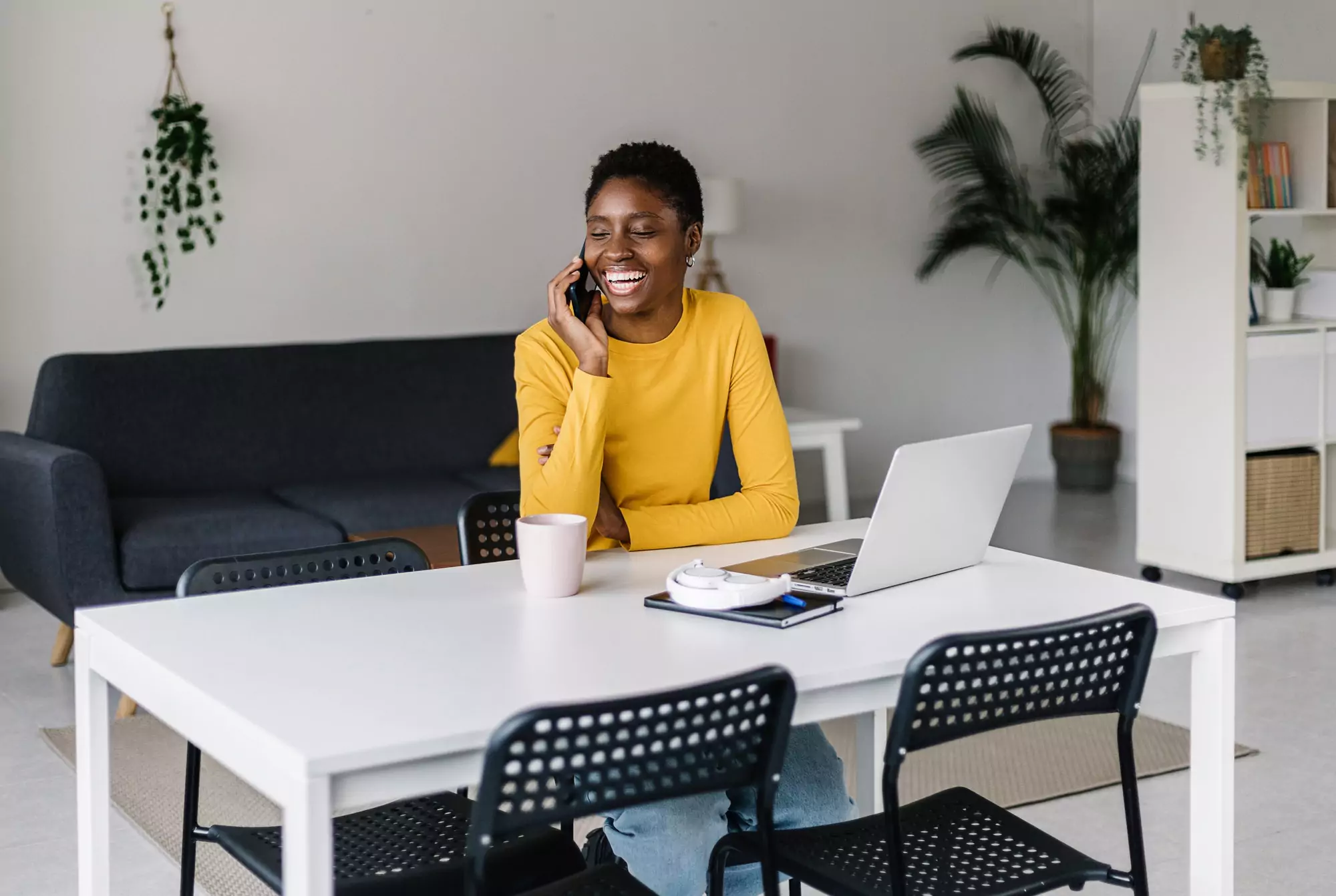 Happy young african woman using mobile phone while working on laptop at home