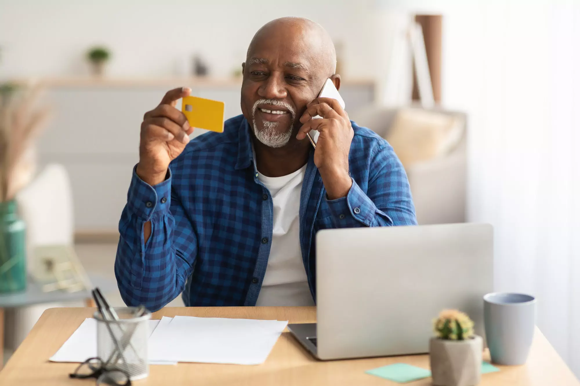 Senior African Man Using Credit Card Talking On Phone Indoors